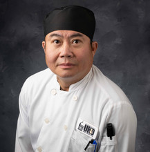 headshot of Justyn Tran wearing chef uniform, pens, and thermometer