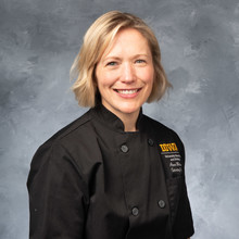 photo of white woman smiling, blue eyes and a blonde bob, wearing black executive chef uniform