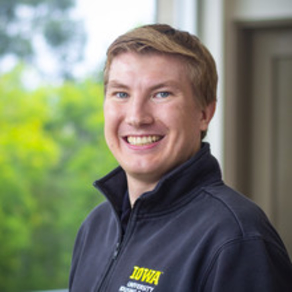 portrait of white man with blue eyes wearing a black quarter-zip against a window with green trees outside