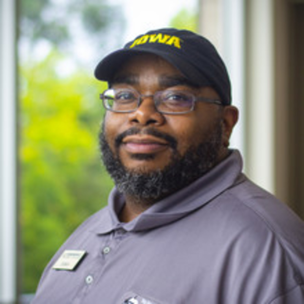 headshot of Edwin West, a black man with a beard wearing glasses, an Iowa cap, and a gray polo shirt