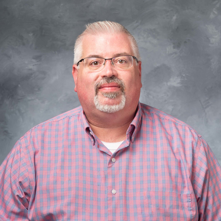portrait of the director of dining, a man wearing glasses with gray hair and a goatee, wearing a pink plaid shirt.