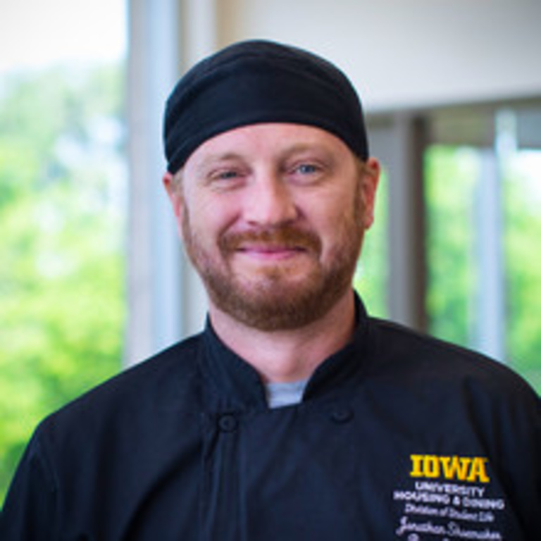 portrait of man in black chef's coat and hat smiling in front of a window