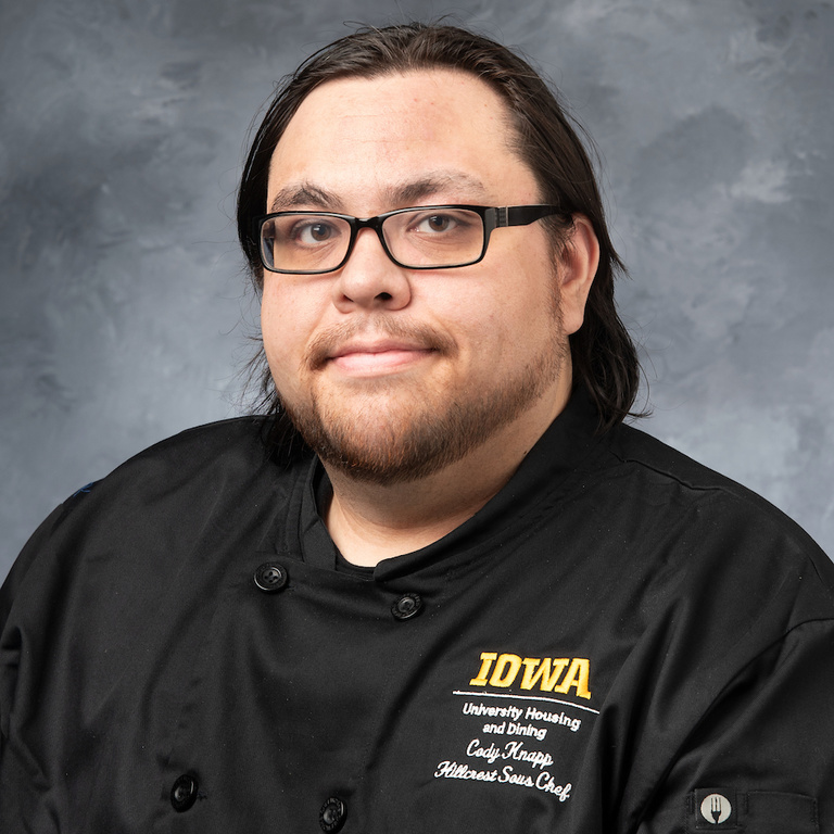 headshot of sous chef Cody Knapp, man with brown hair and beard, wearing rectangular framed glasses and black chef's coat